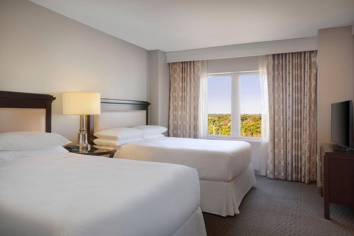A hotel room with two beds, a bedside table with a lamp, a TV, and a large window with curtains.