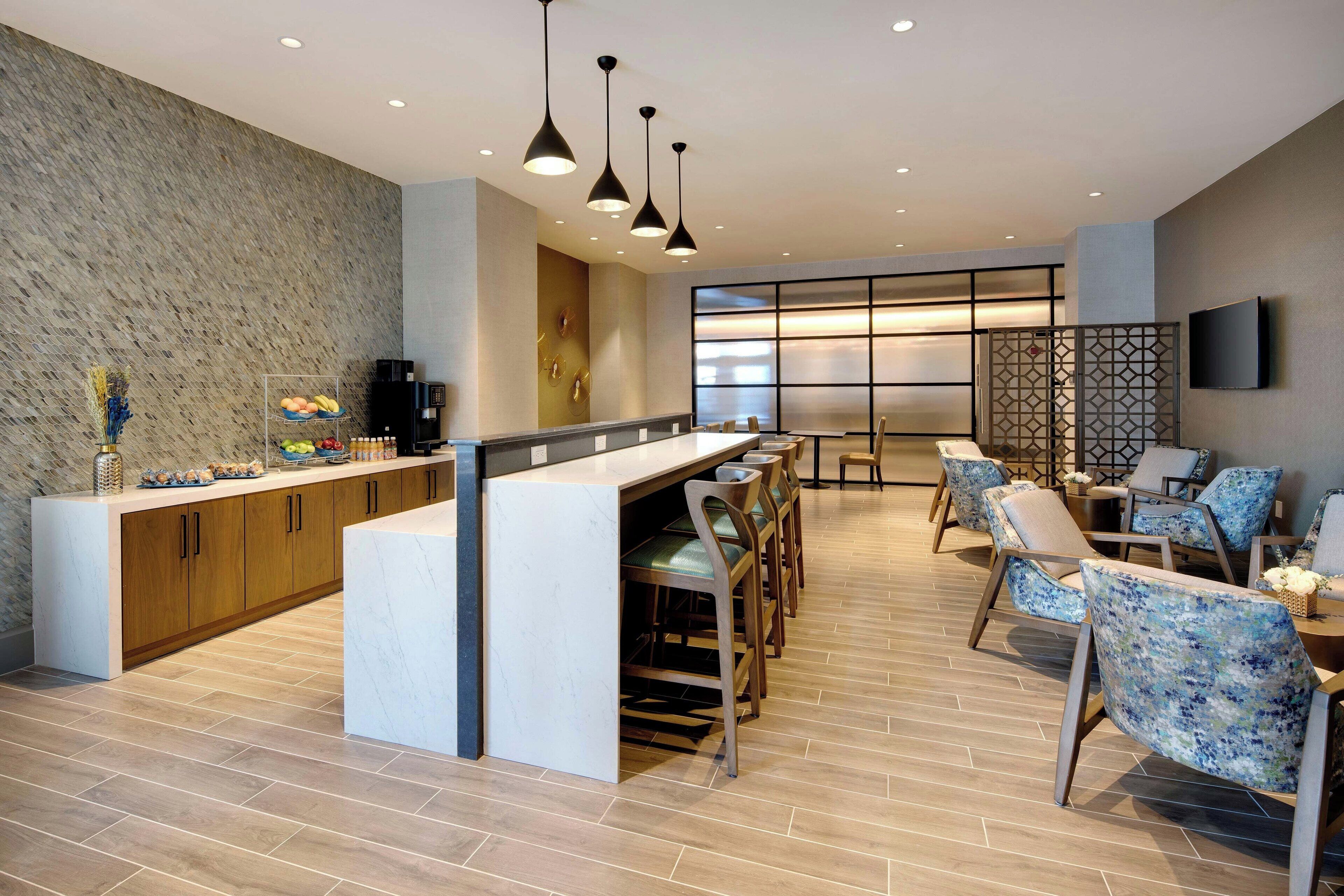 Uma área de bar moderna com bancada de mármore, armários de madeira e banquetas. Há luminárias pendentes acima da bancada e um espaço de estar com cadeiras estofadas.
