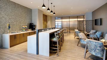 Uma área de bar moderna com bancada de mármore, armários de madeira e banquetas. Há luminárias pendentes acima da bancada e um espaço de estar com cadeiras estofadas.