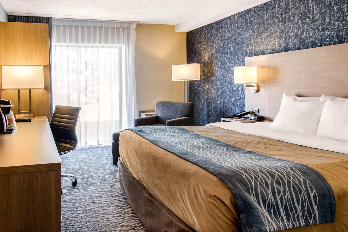 Habitación de hotel con una cama grande, un escritorio con lámpara, una silla y vistas al exterior a través de una ventana.