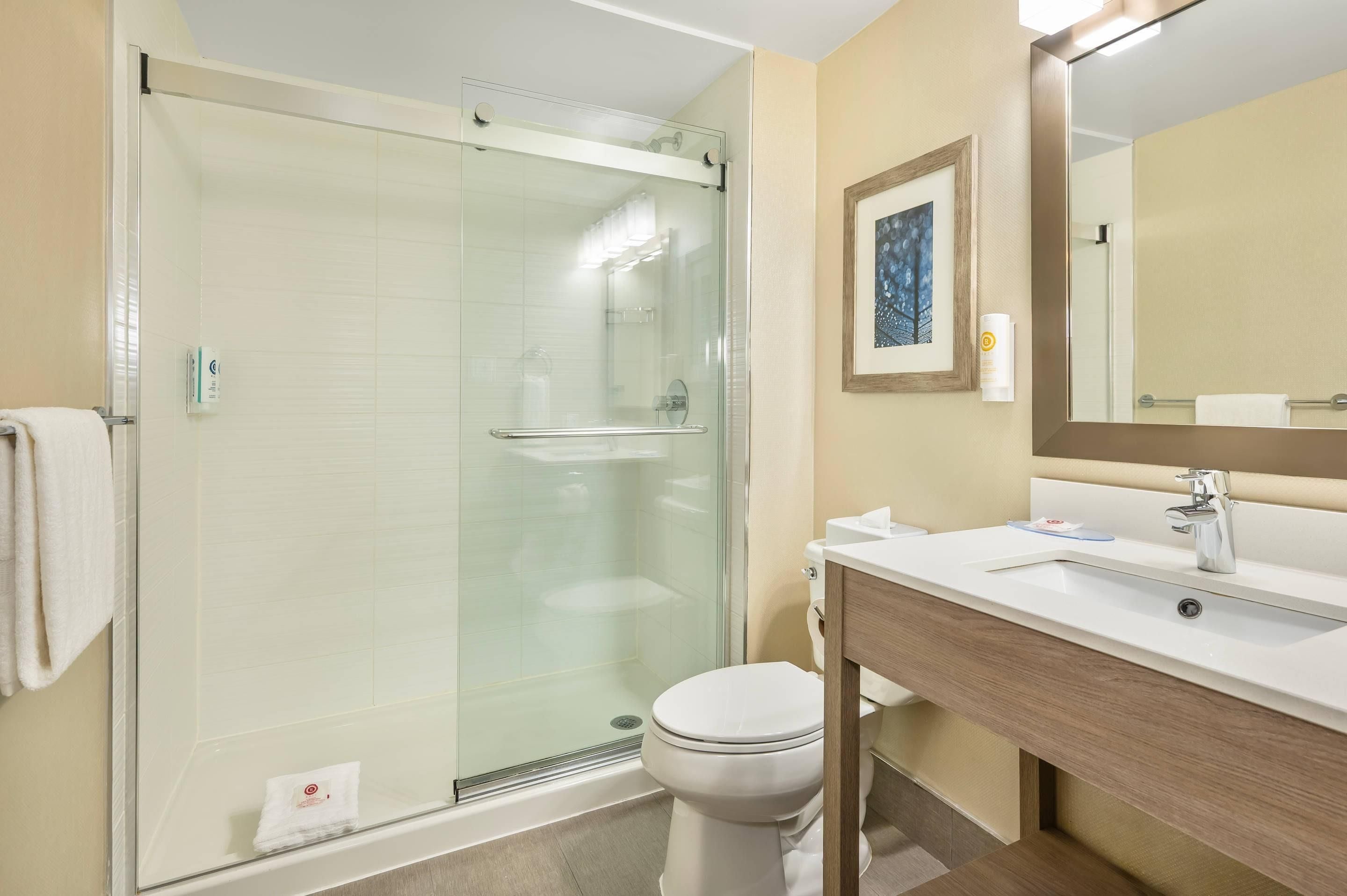 Un baño moderno con ducha envuelta en vidrio, inodoro blanco y tocador de madera con espejo.
