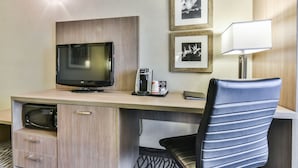 A hotel room with a wooden desk, a flat-screen TV, a coffee maker, a lamp, and framed pictures on the wall.