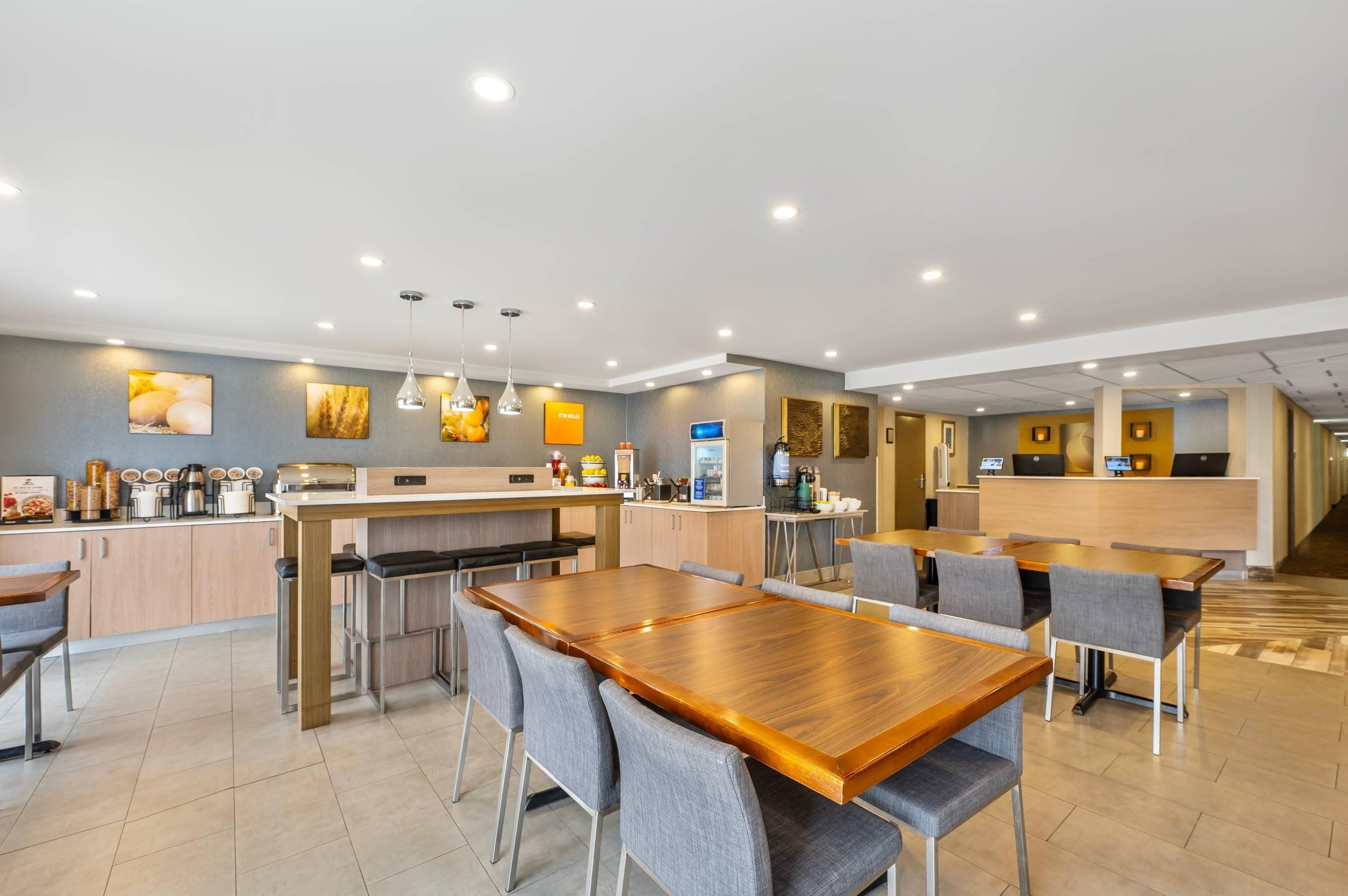 A modern hotel dining area with a wooden table and grey upholstered chairs, a kitchen area with a bar, and a reception desk.