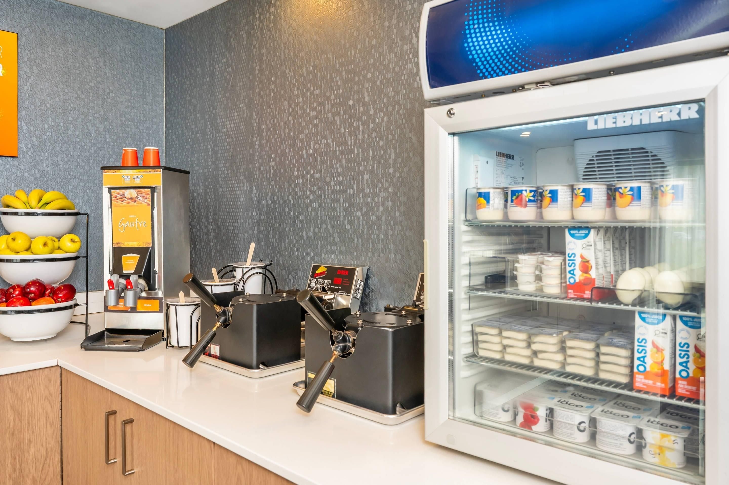 A self-service beverage station with coffee machines and a refrigerator stocked with milk and juice cartons.