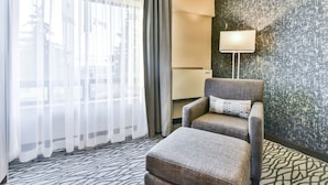 A hotel room with a grey armchair, a matching ottoman, a patterned rug, a large window with sheer curtains, and a wall-mounted lamp.