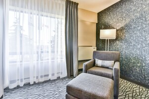 A hotel room with a grey armchair, a matching ottoman, a patterned rug, a large window with sheer curtains, and a wall-mounted lamp.