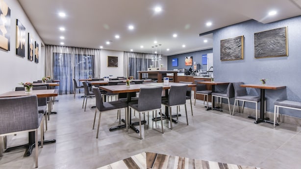 A modern hotel dining area with multiple tables and chairs, a kitchen in the background, and artwork on the walls.