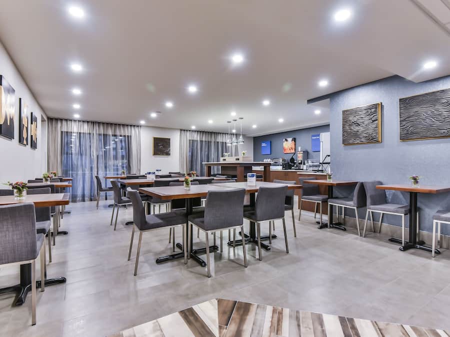 A modern hotel dining area with multiple tables and chairs, a kitchen in the background, and artwork on the walls.