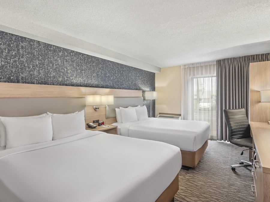 Quarto de hotel com duas camas, uma escrivaninha, uma cadeira e uma janela grande com cortinas.