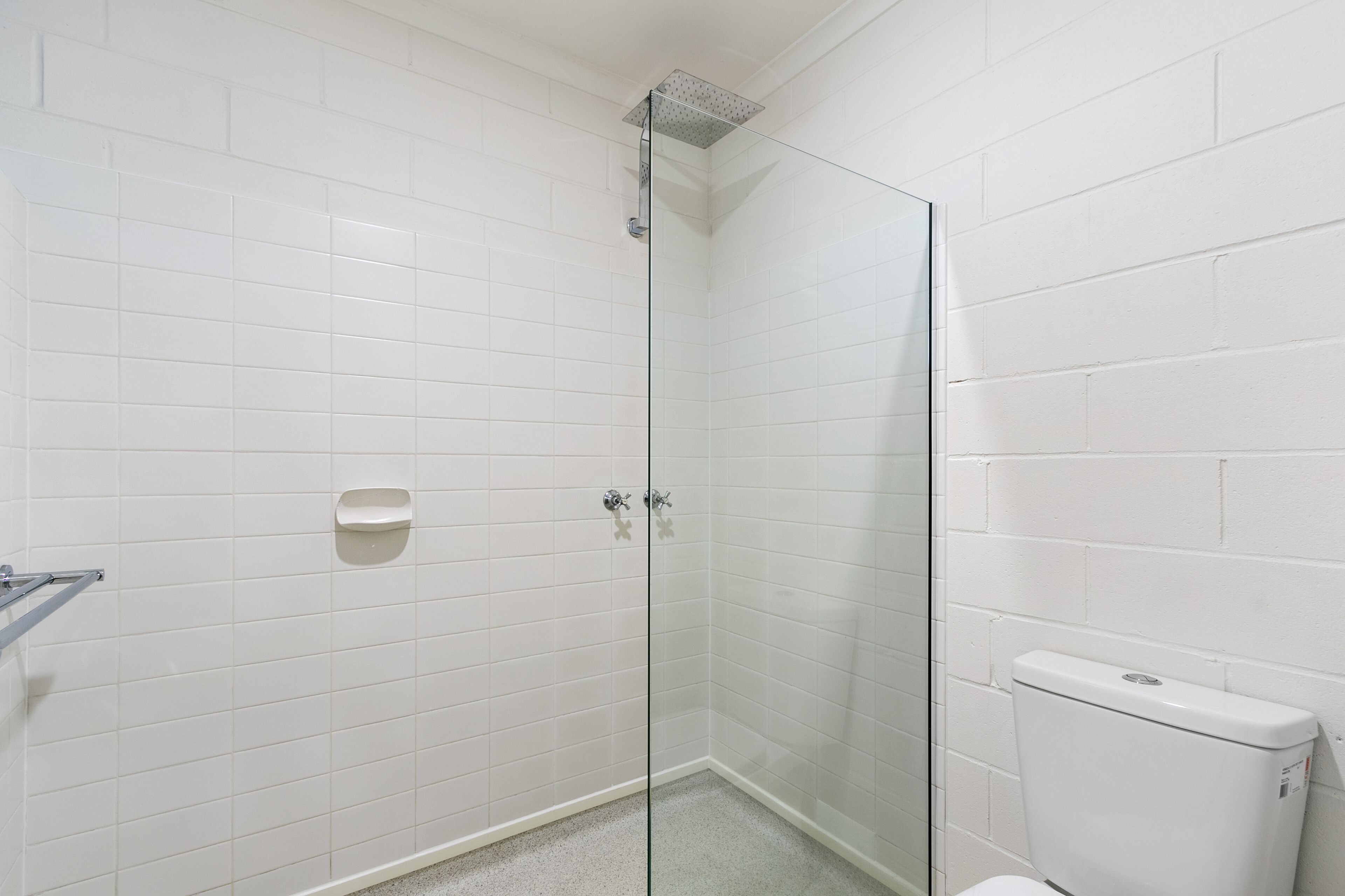 Un baño con paredes de azulejos blancos, cabina de ducha de vidrio y váter blanco.