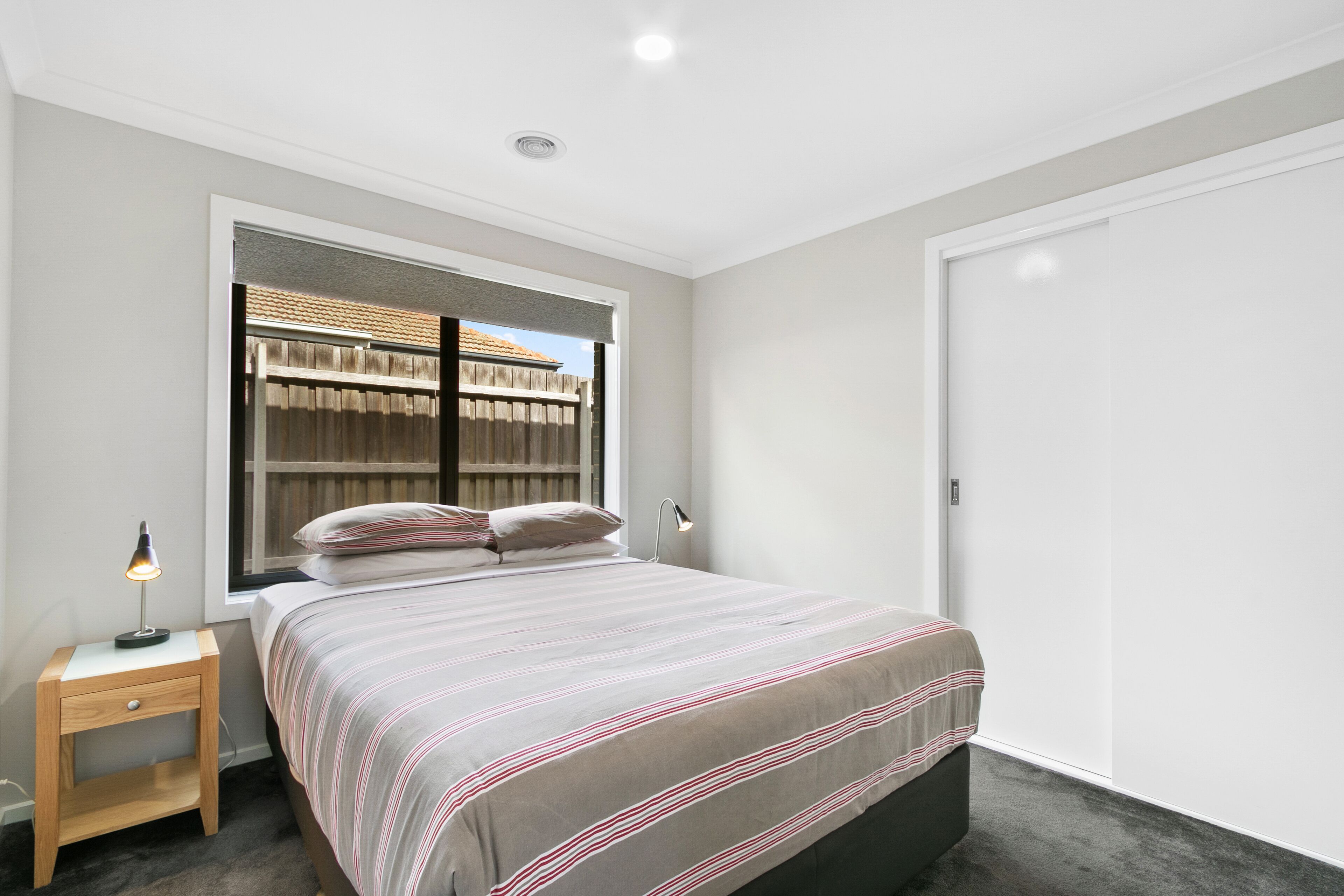 A bedroom with a bed, bedside lamps, a window with a view of a fence, and a wardrobe.