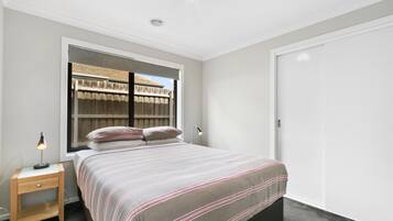 A bedroom with a bed, bedside lamps, a window with a view of a fence, and a wardrobe.
