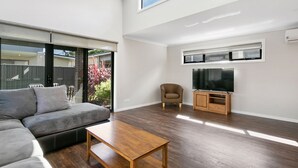 A modern living room with a sectional sofa, a wooden coffee table, a television on a wooden stand, and a sliding glass door leading to an outdoor area. - The Aspen & Apartments (Sale)