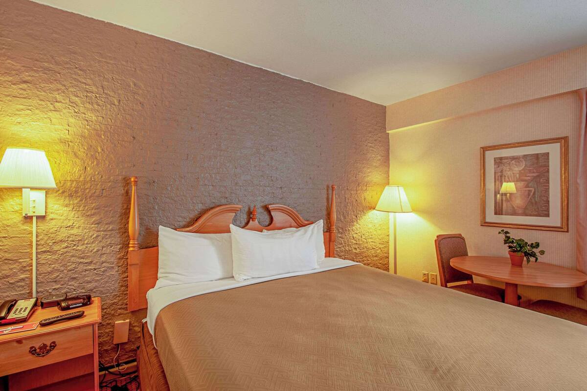 A hotel room with a large bed, bedside lamps, a wooden nightstand, a table with a chair, and a framed picture on the wall.