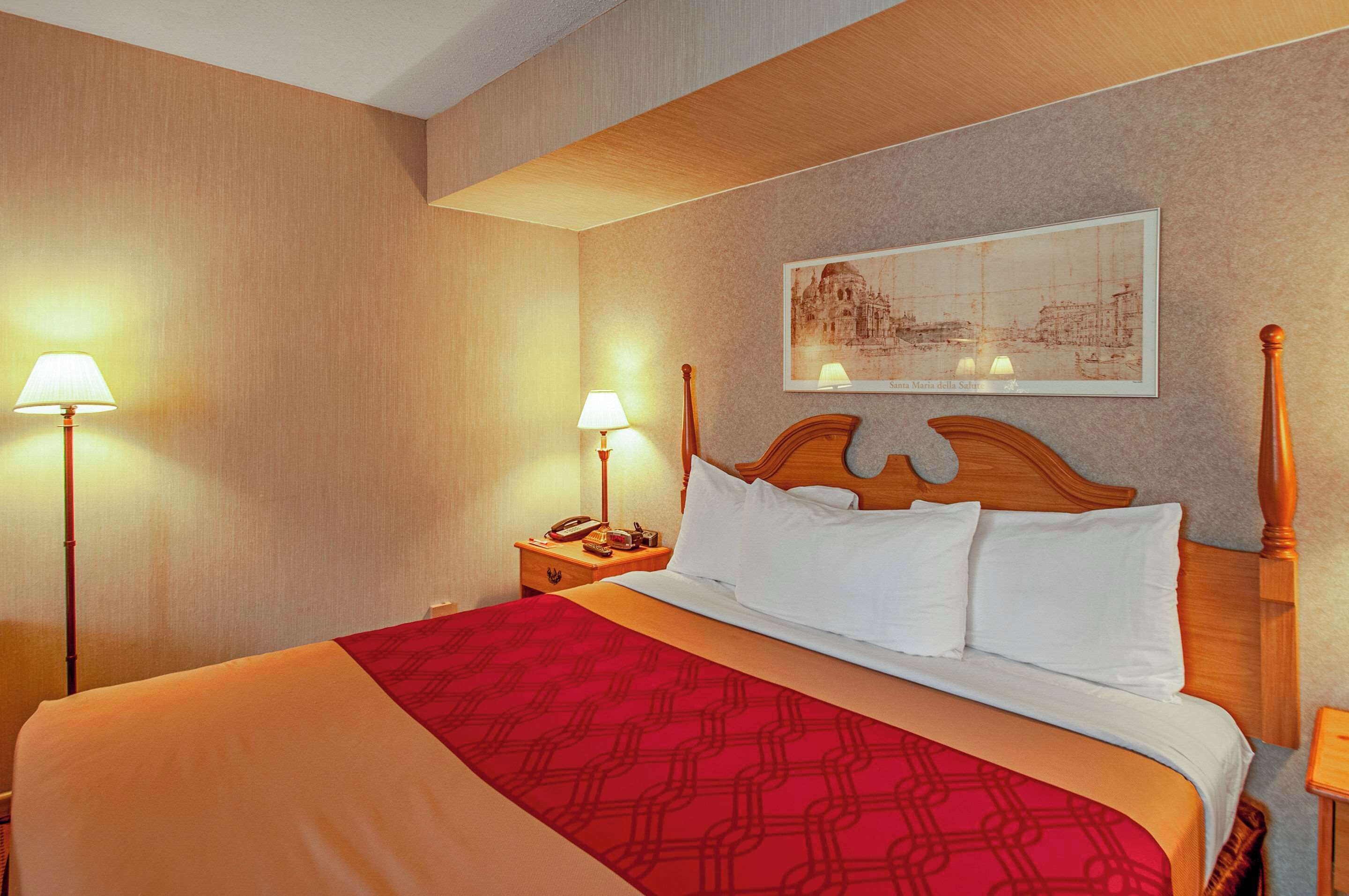 A hotel room with a large bed, two bedside lamps, a wooden headboard, a red and white bedspread, and a framed picture on the wall.