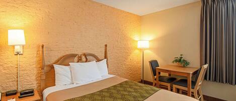 Una habitación de hotel con cama, mesitas de noche, un escritorio con silla, lámpara y planta.
