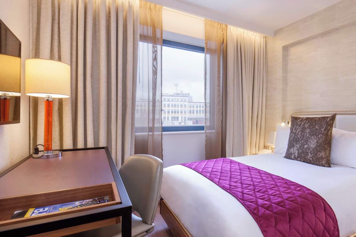 A hotel room with a bed, a desk, a chair, a lamp, and a window with curtains.