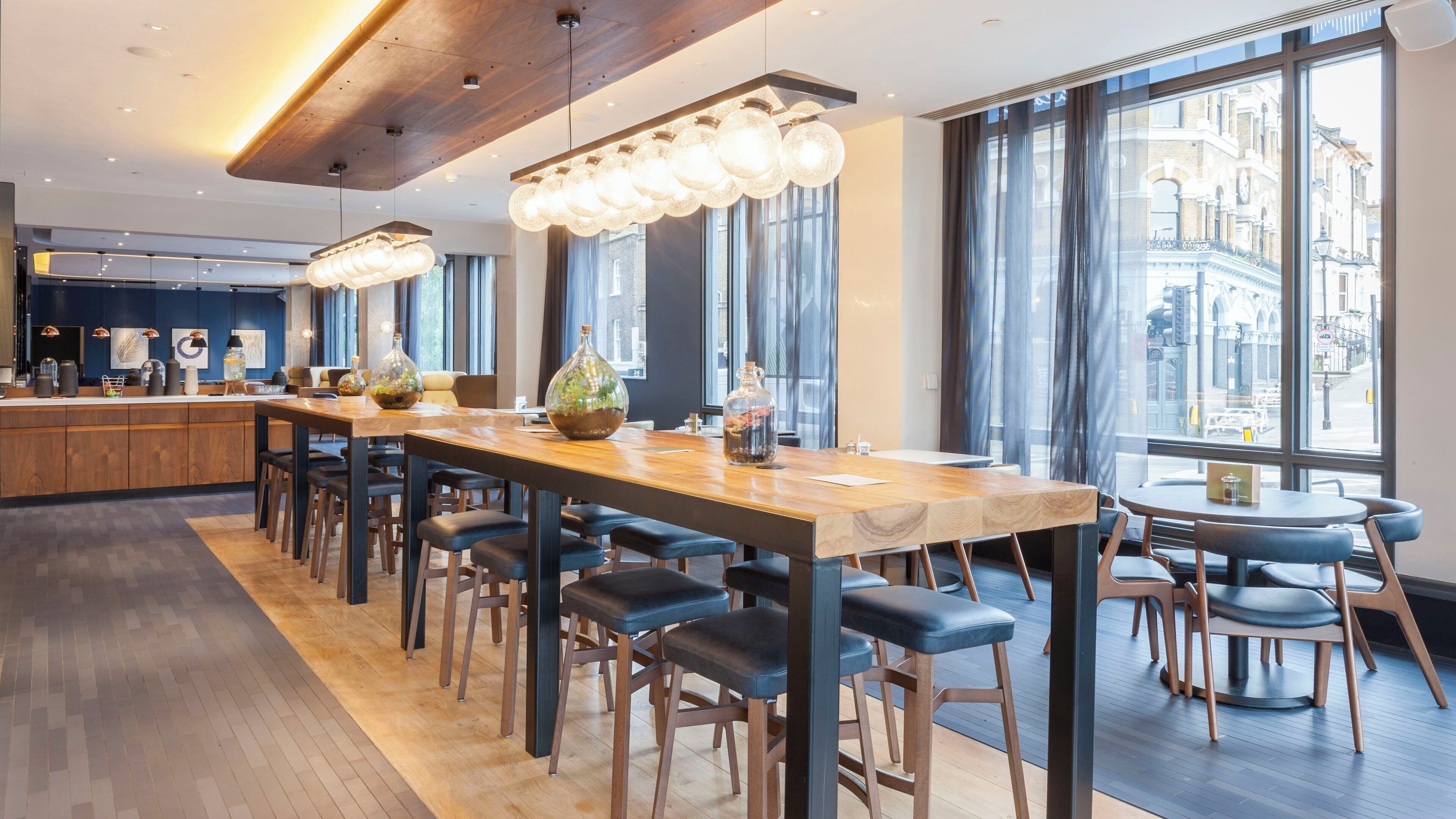 A modern bar area with a long wooden counter, multiple stools, and hanging light fixtures.