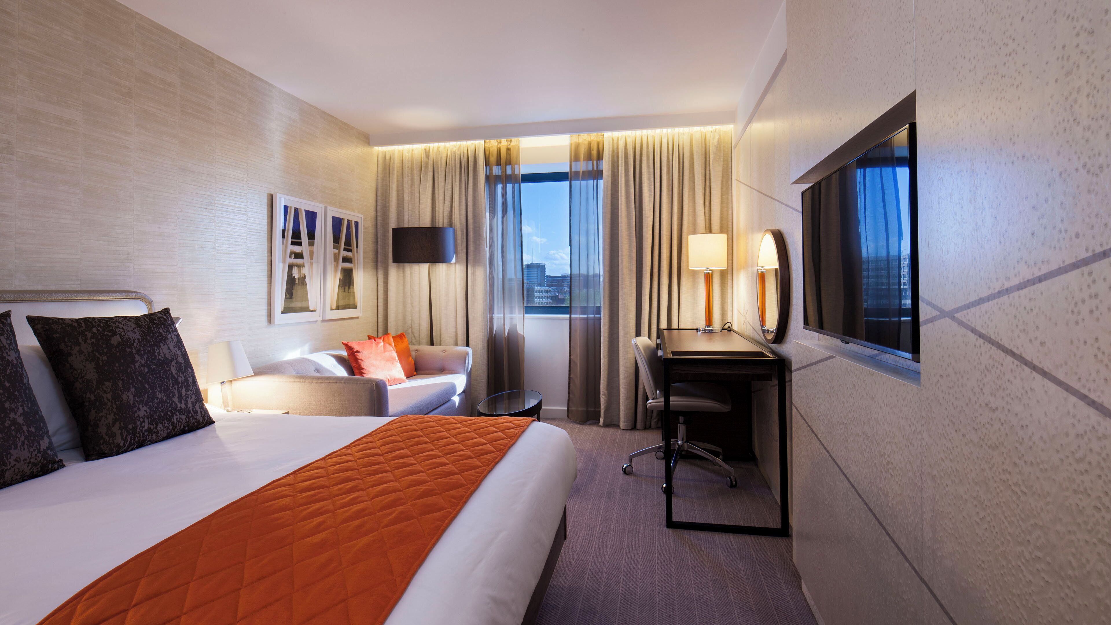 A hotel room with a bed, a desk, a chair, a sofa, and a large window with curtains.
