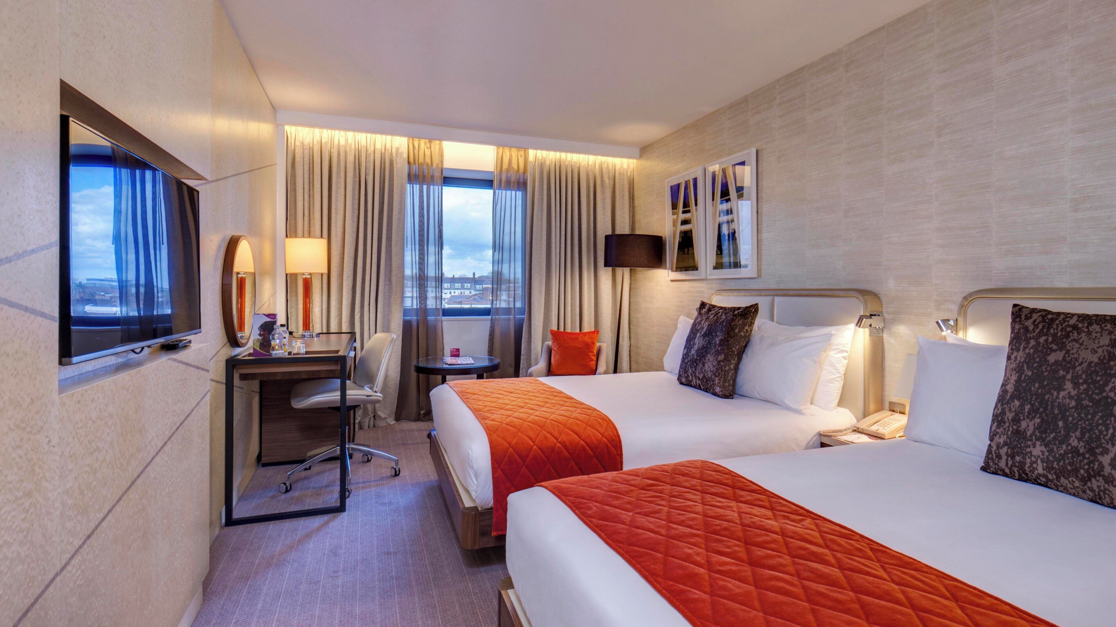 A hotel room with two beds, a desk with a chair, a TV, and a window with curtains.