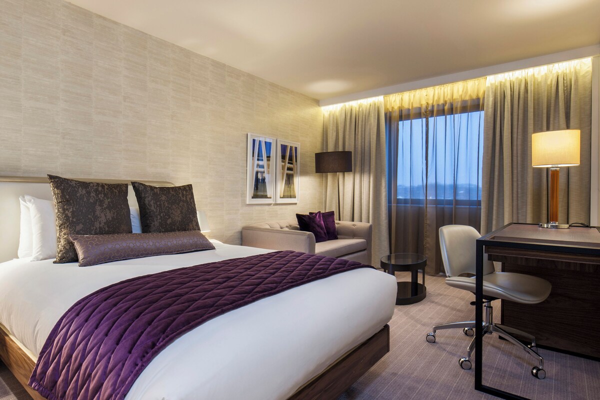 A hotel room with a large bed, a desk, a chair, and a view of the city.