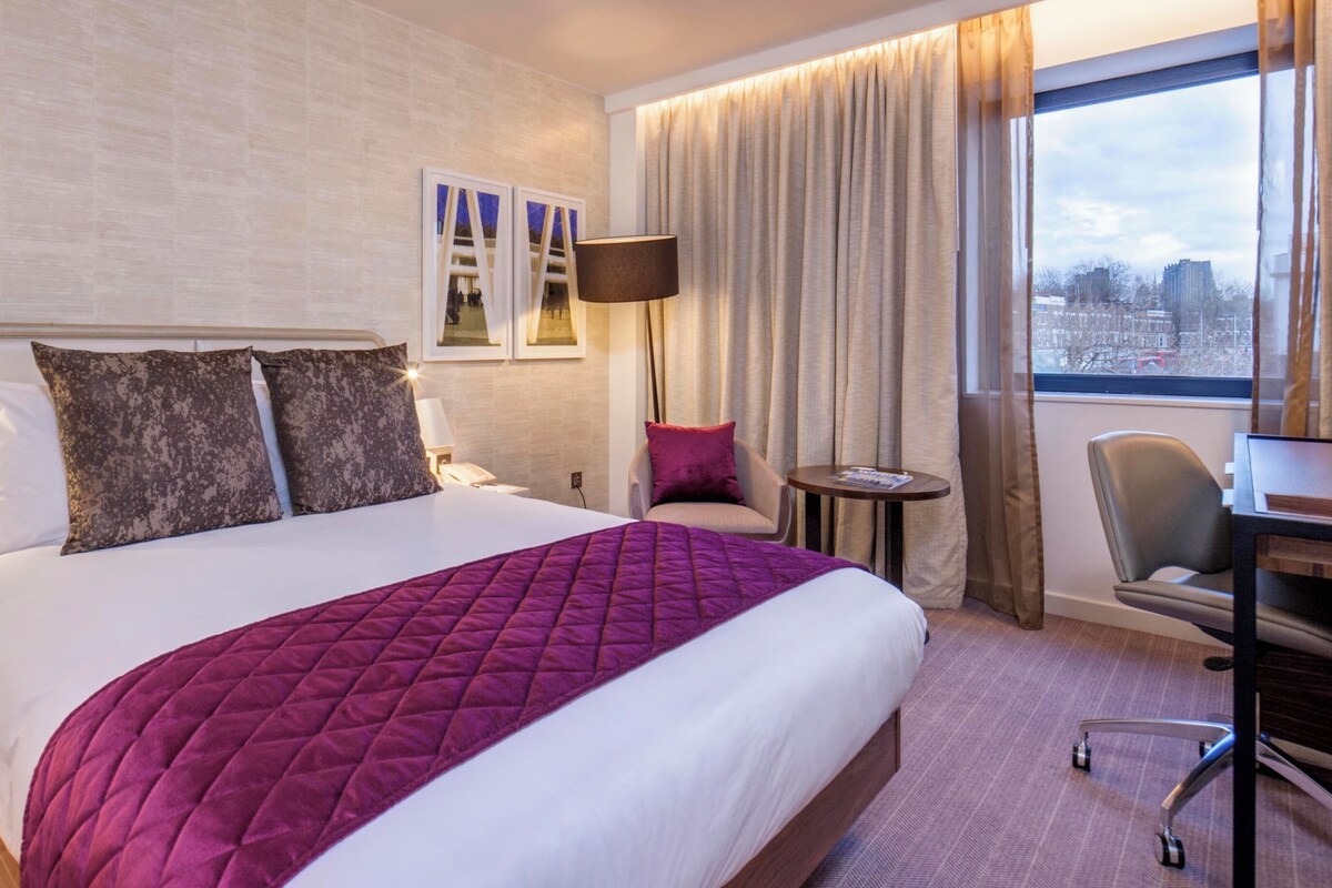 A hotel room with a large bed, a desk, a chair, and a view of the city through the window.