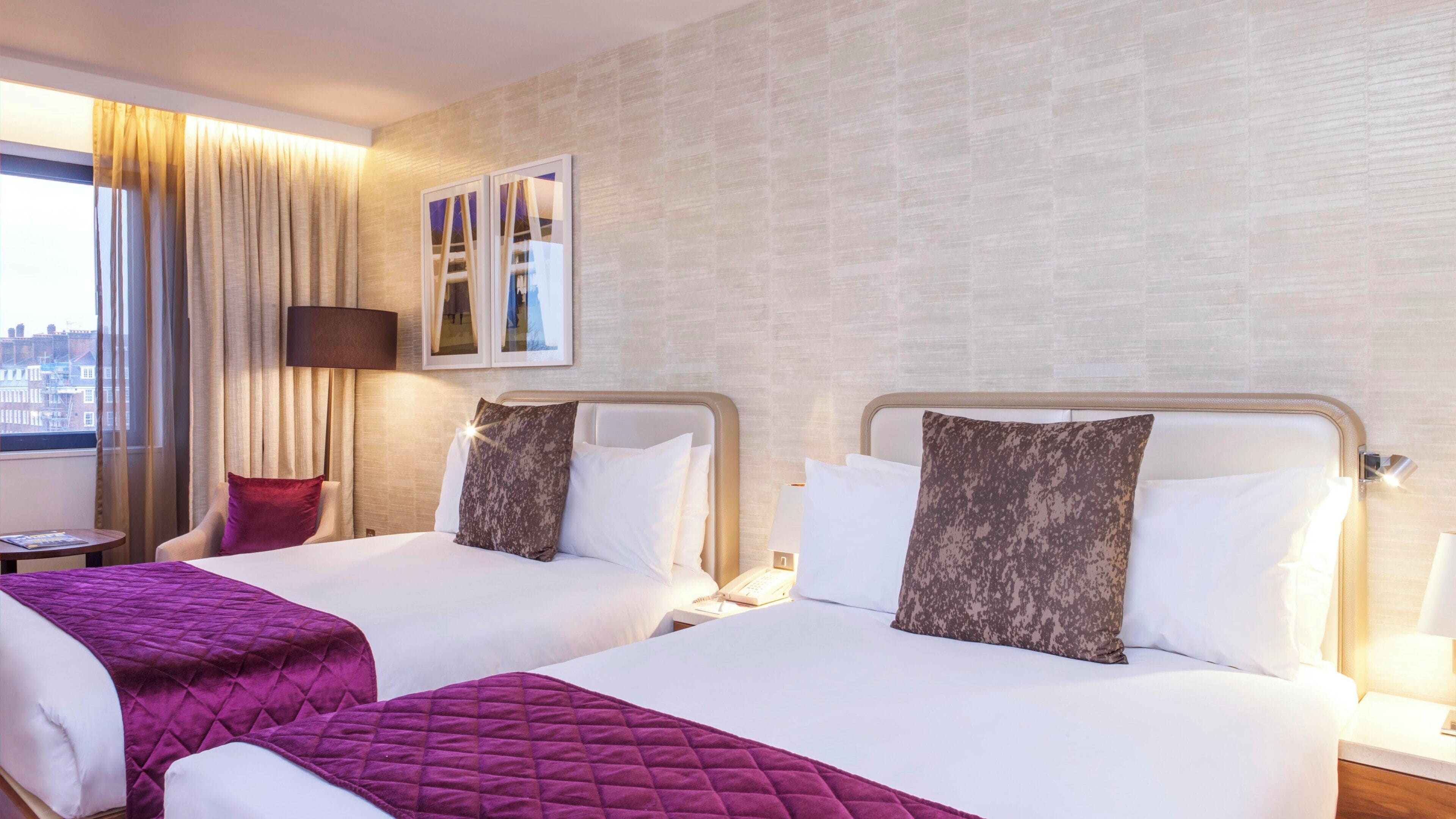 A hotel room with two beds, a city view, a window with curtains, and a small table with a lamp.