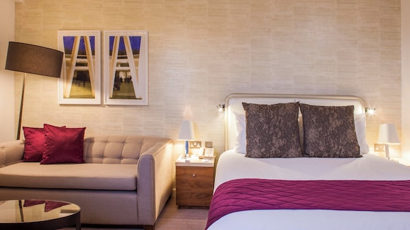 A hotel room with a bed, a sofa, a nightstand, a lamp, and two framed pictures on the wall.