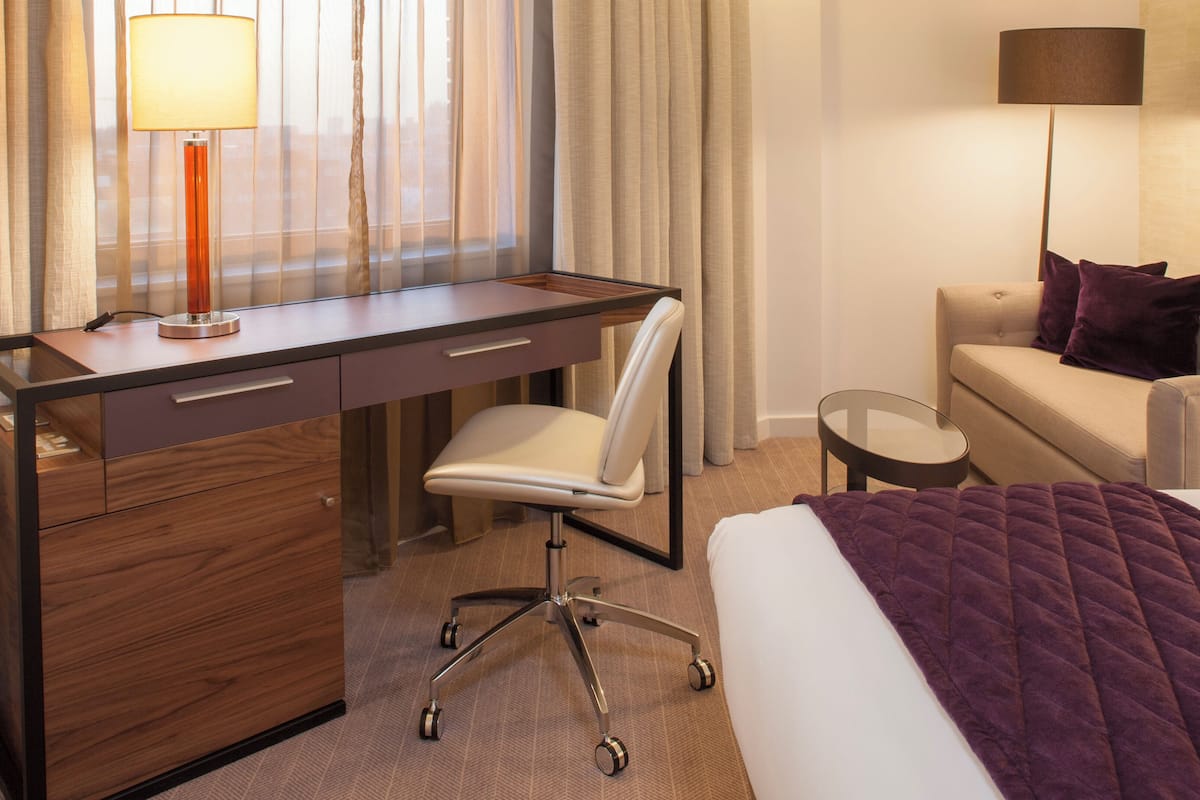 A hotel room with a wooden desk, a white chair, a bed with a purple blanket, a small round table, and a lamp.