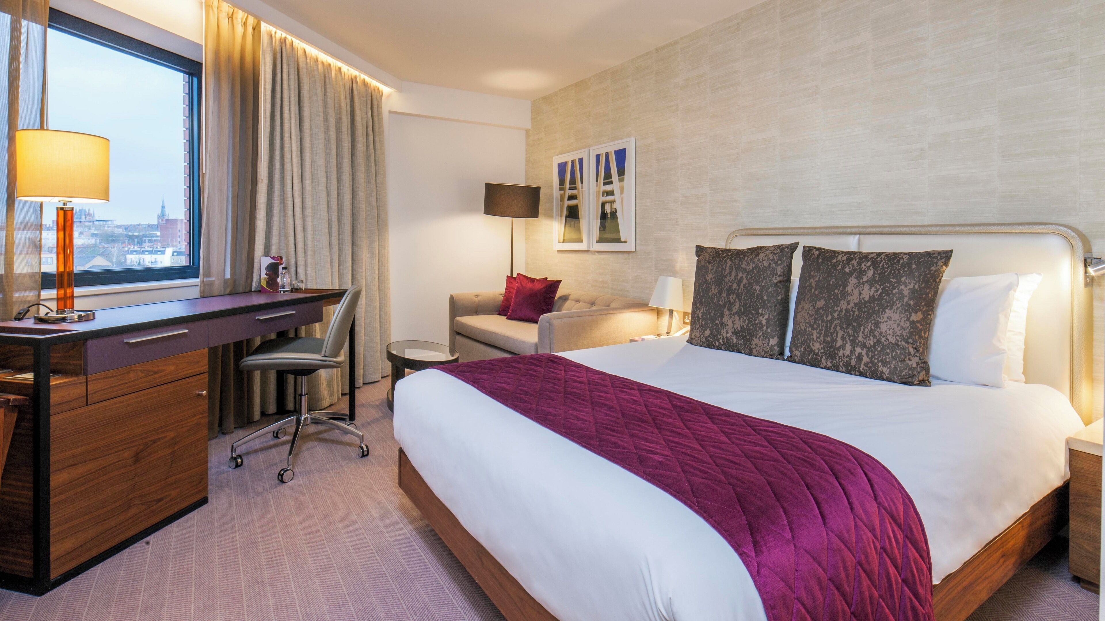 Quarto de hotel com cama, escrivaninha, cadeira, sofá e vista para a cidade pela janela.
