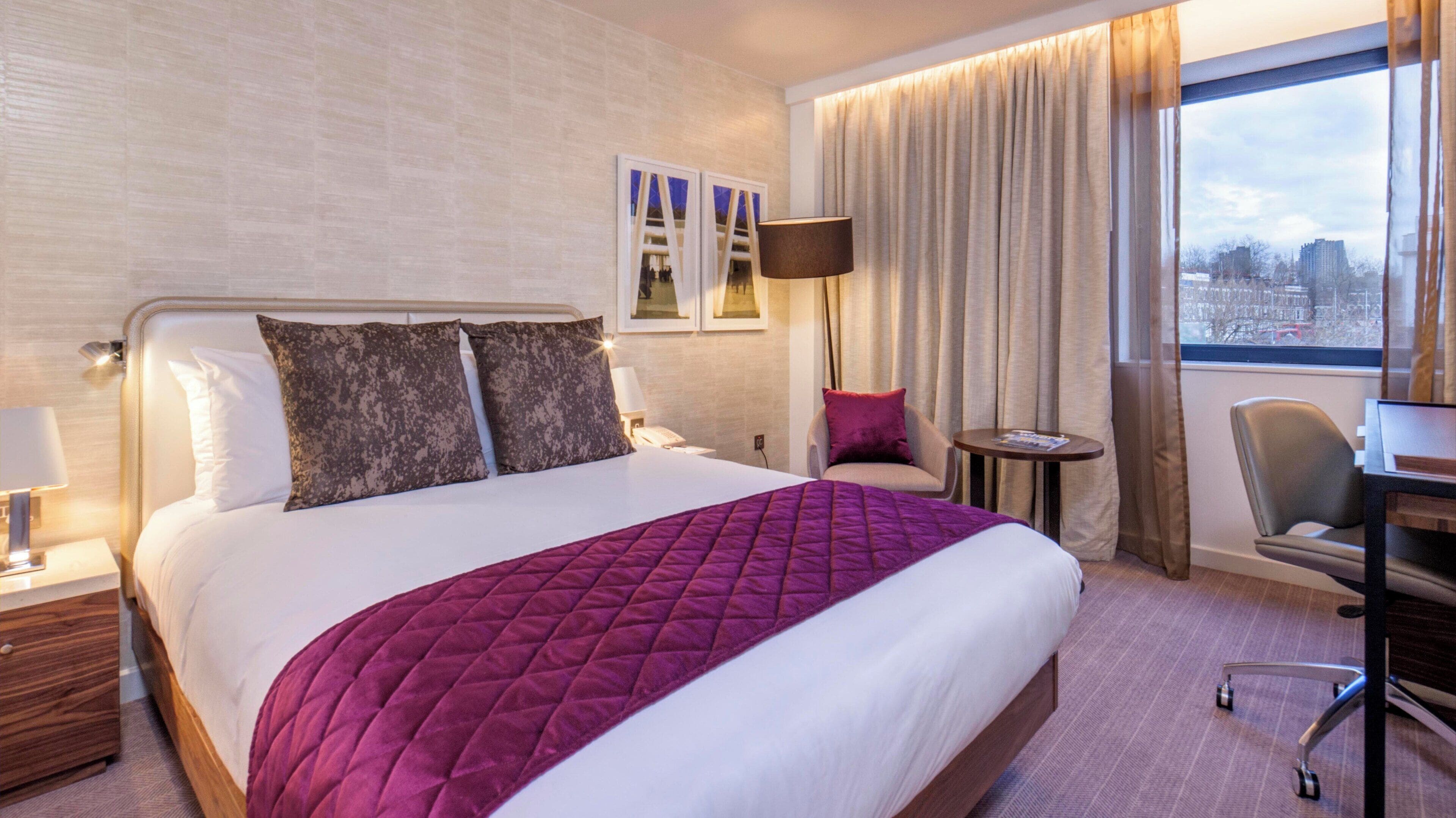A hotel room with a bed, a desk, a chair, a lamp, and a view of the city through the window.
