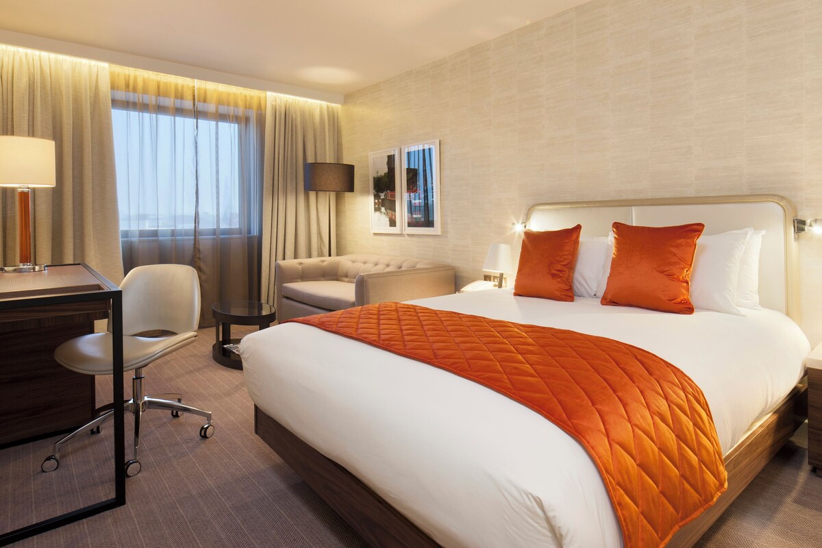 A hotel room with a large bed, a desk, a chair, a sofa, and a nightstand with a lamp.