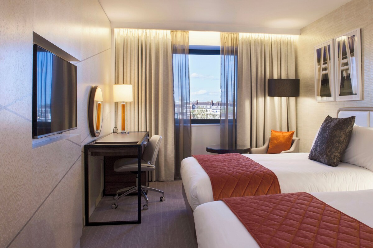 A hotel room with two beds, a desk with a chair, a window with curtains, and a view of buildings.