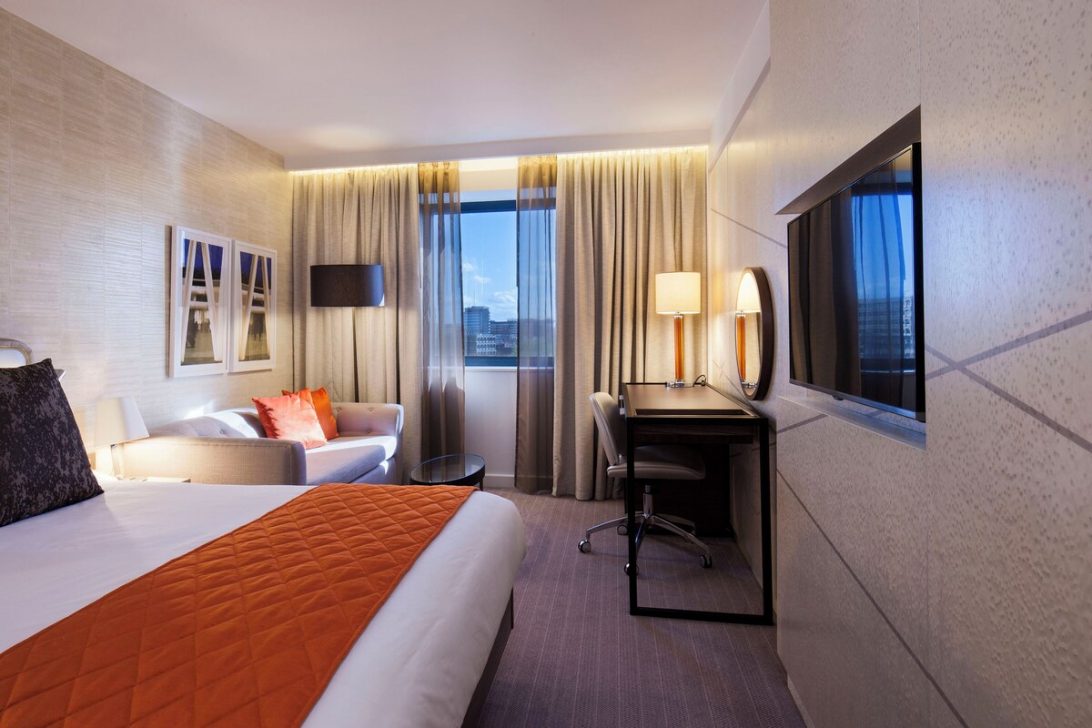 A hotel room with a large bed, a desk, a chair, a sofa, a TV, and a window with curtains.