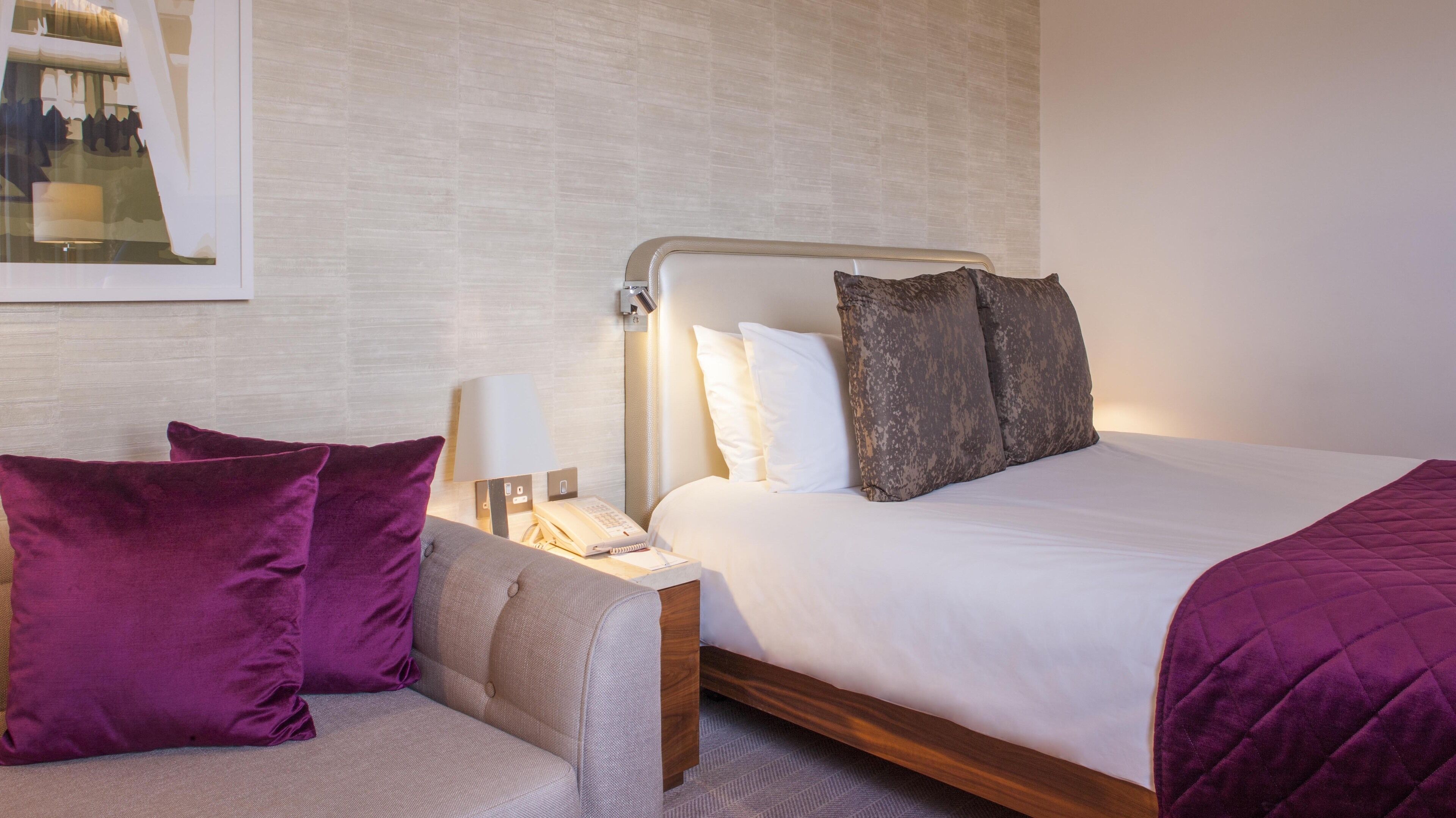 A hotel room with a bed, a nightstand with a lamp, a chair, and a painting on the wall.
