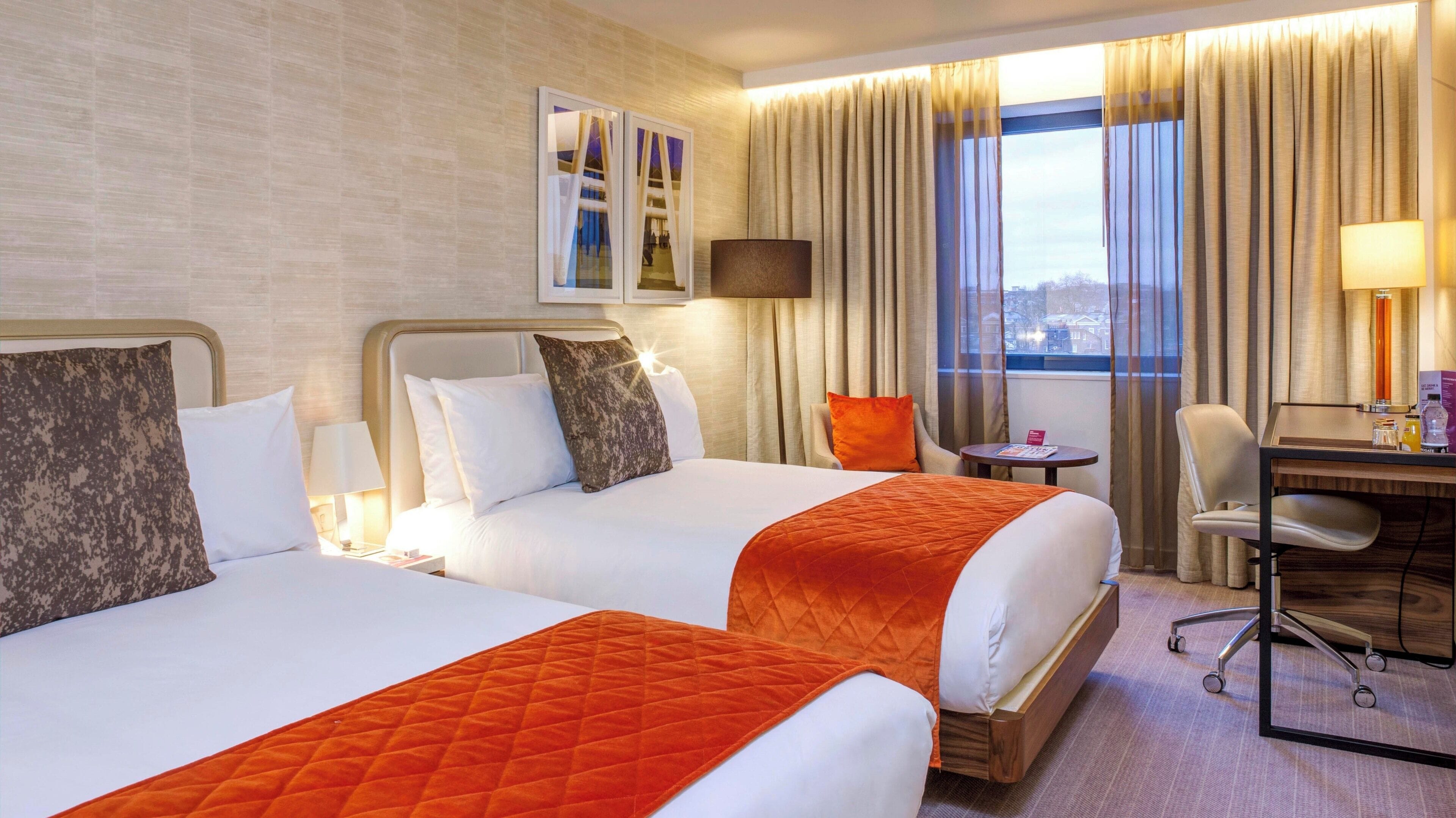 Een hotelkamer met twee bedden, een bureau, een stoel, een lamp en een raam met gordijnen.