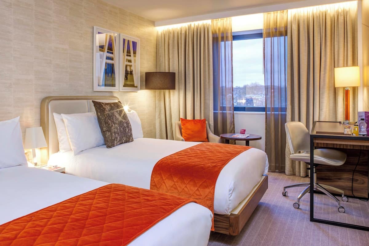 A hotel room with two beds, a desk, a chair, a lamp, and a window with curtains.