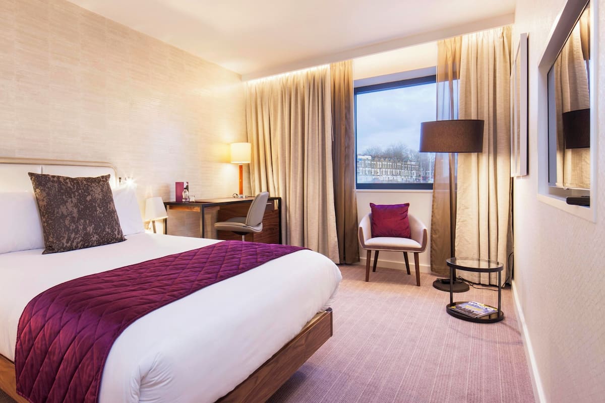 A hotel room with a large bed, a desk, a chair, a small table, and a window with curtains.