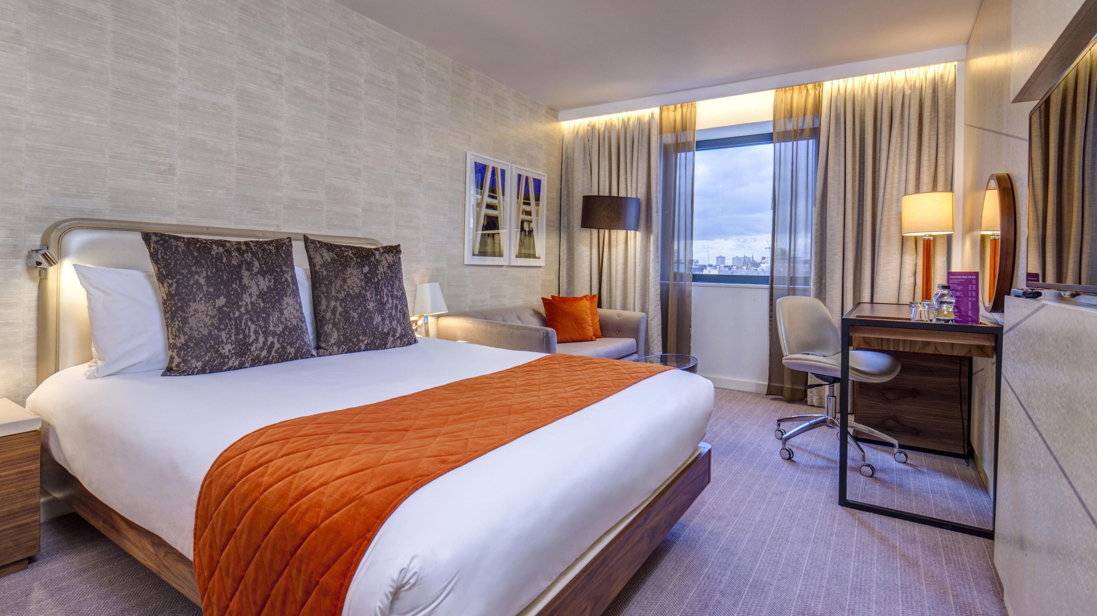 A hotel room with a large bed, a desk, a chair, and a view of the city through the window.
