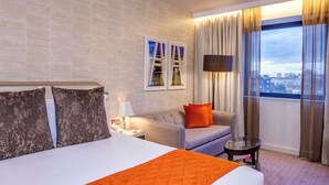 Habitación de hotel con una cama grande, un sofá, una mesita y vistas a la ciudad desde la ventana.