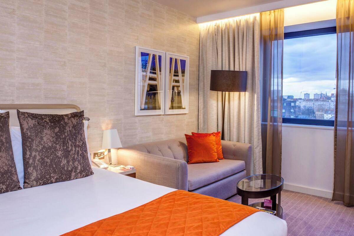A hotel room with a large bed, a sofa, a small table, and a view of the city through the window.