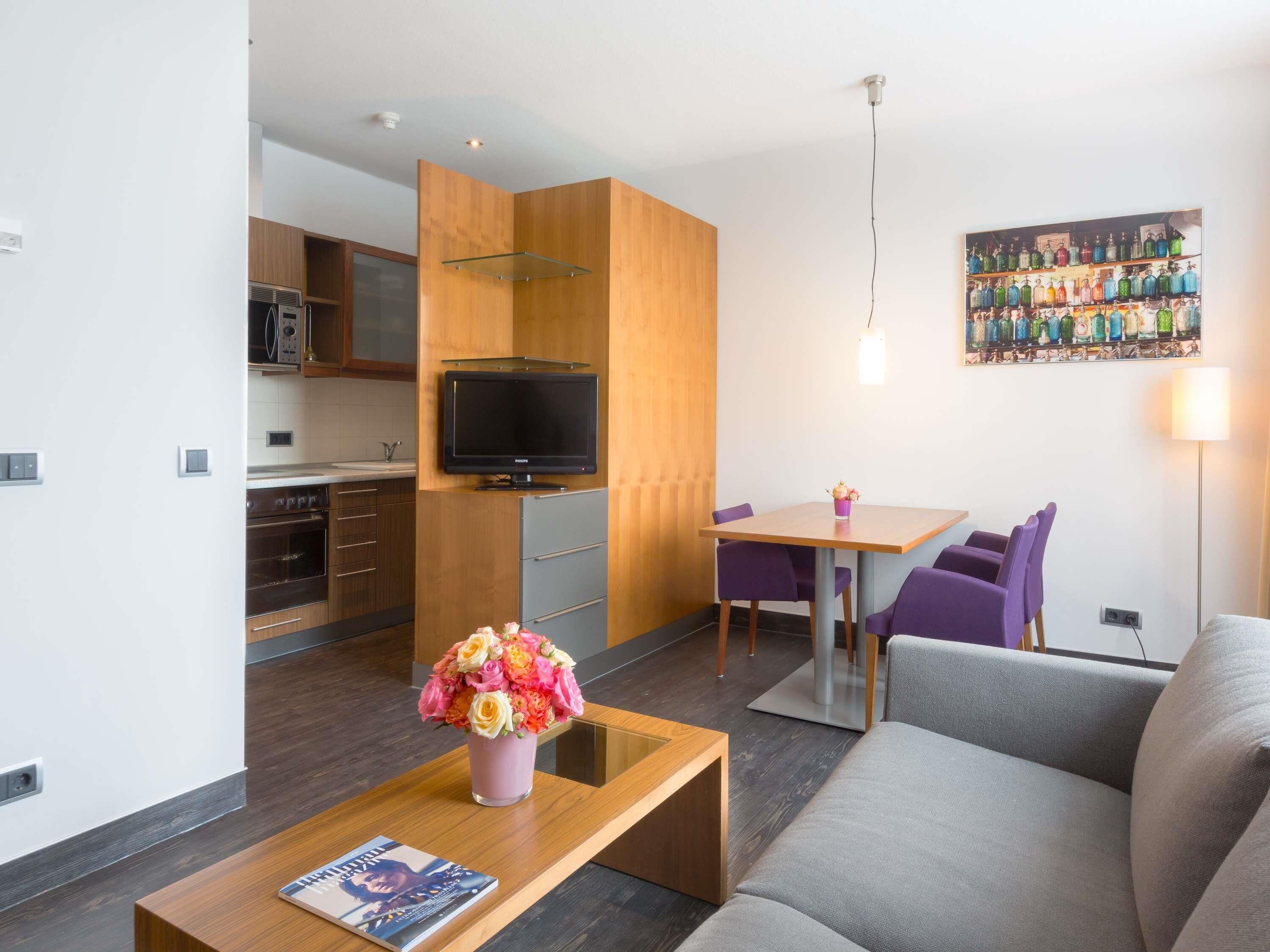 Un salon moderne avec un canapé, une table basse et un téléviseur. On y trouve également un coin repas avec une table et des chaises, et une cuisine à l’arrière-plan.