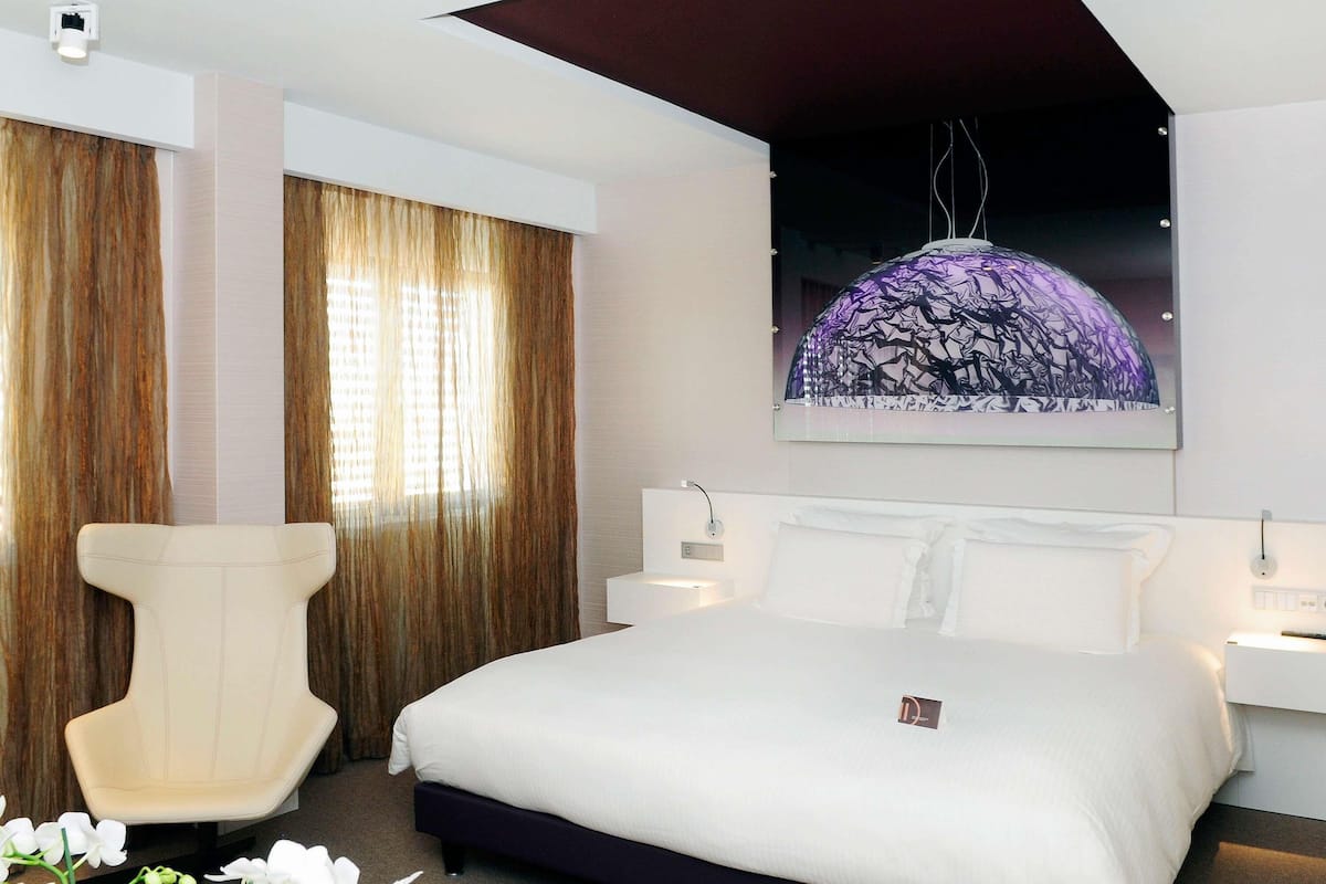 A hotel room with a large bed, a chair, a nightstand, and a wall-mounted artwork.