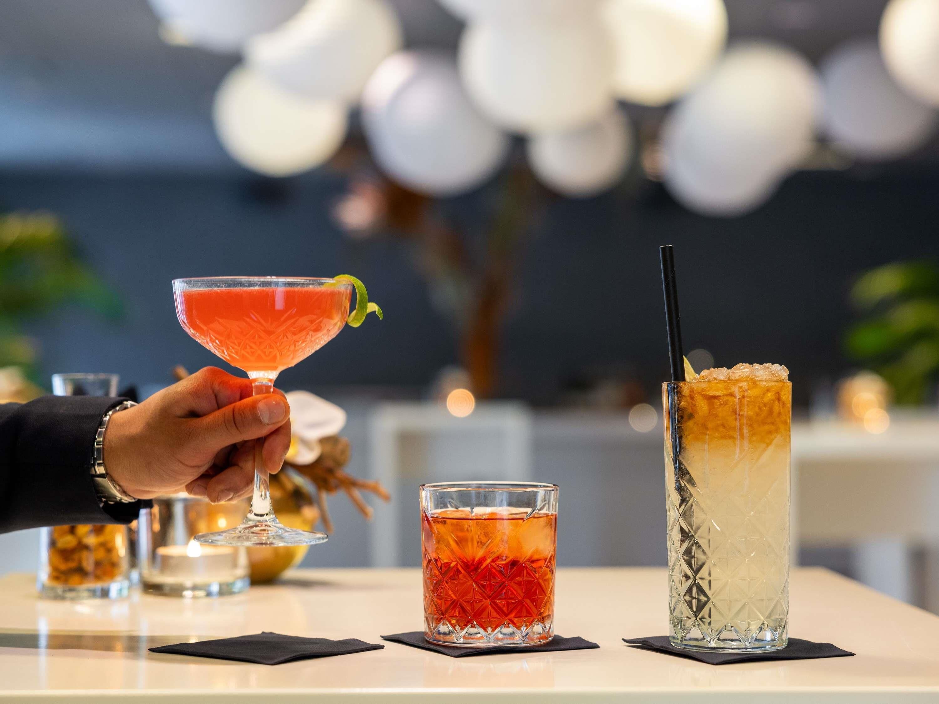 A bar setting with a hand holding a cocktail glass, two other glasses on the table, and a candle in the background.