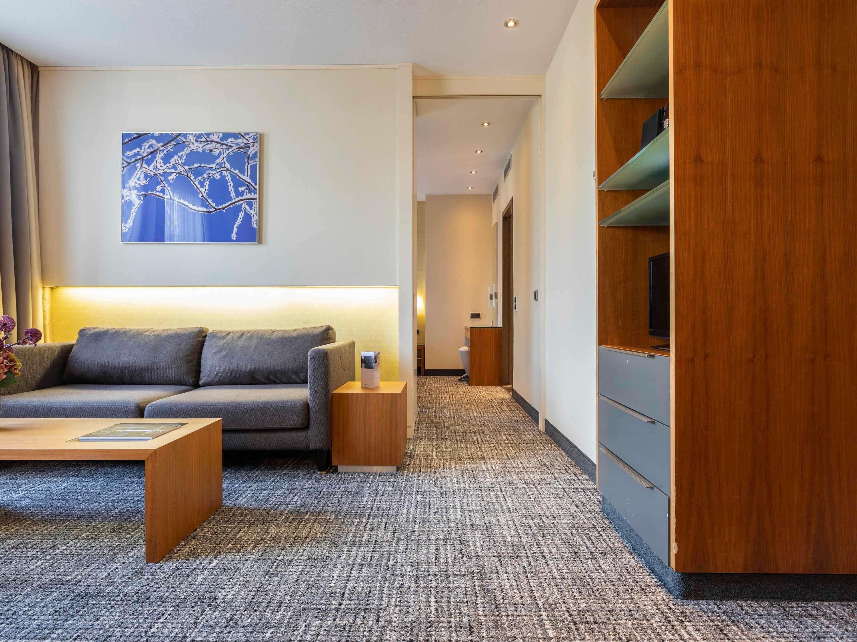 A modern hotel room with a grey sofa, a coffee table, a wooden TV stand, and a carpeted floor.