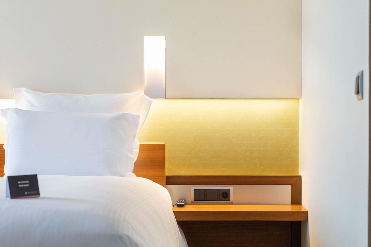 A neatly made bed with white linens, a bedside table with a lamp, and a small electronic device on the table.