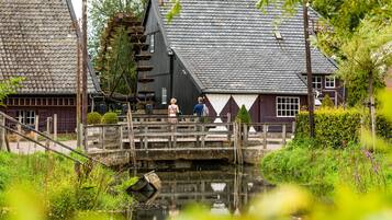 A traditional Dutch house with a steeply pitched roof, a wooden bridge over a canal, and lush greenery surrounding the property.