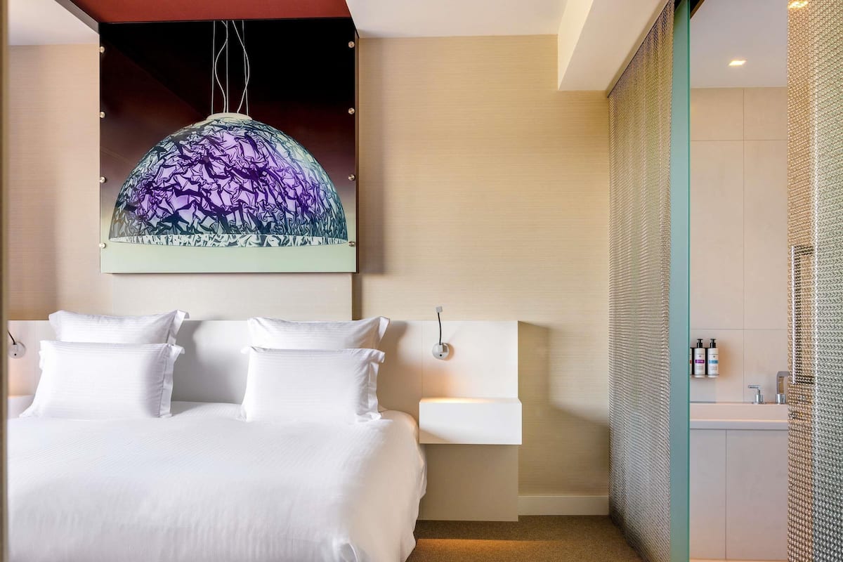 A modern hotel room with a large bed, a wall-mounted sink, and a shower area.
