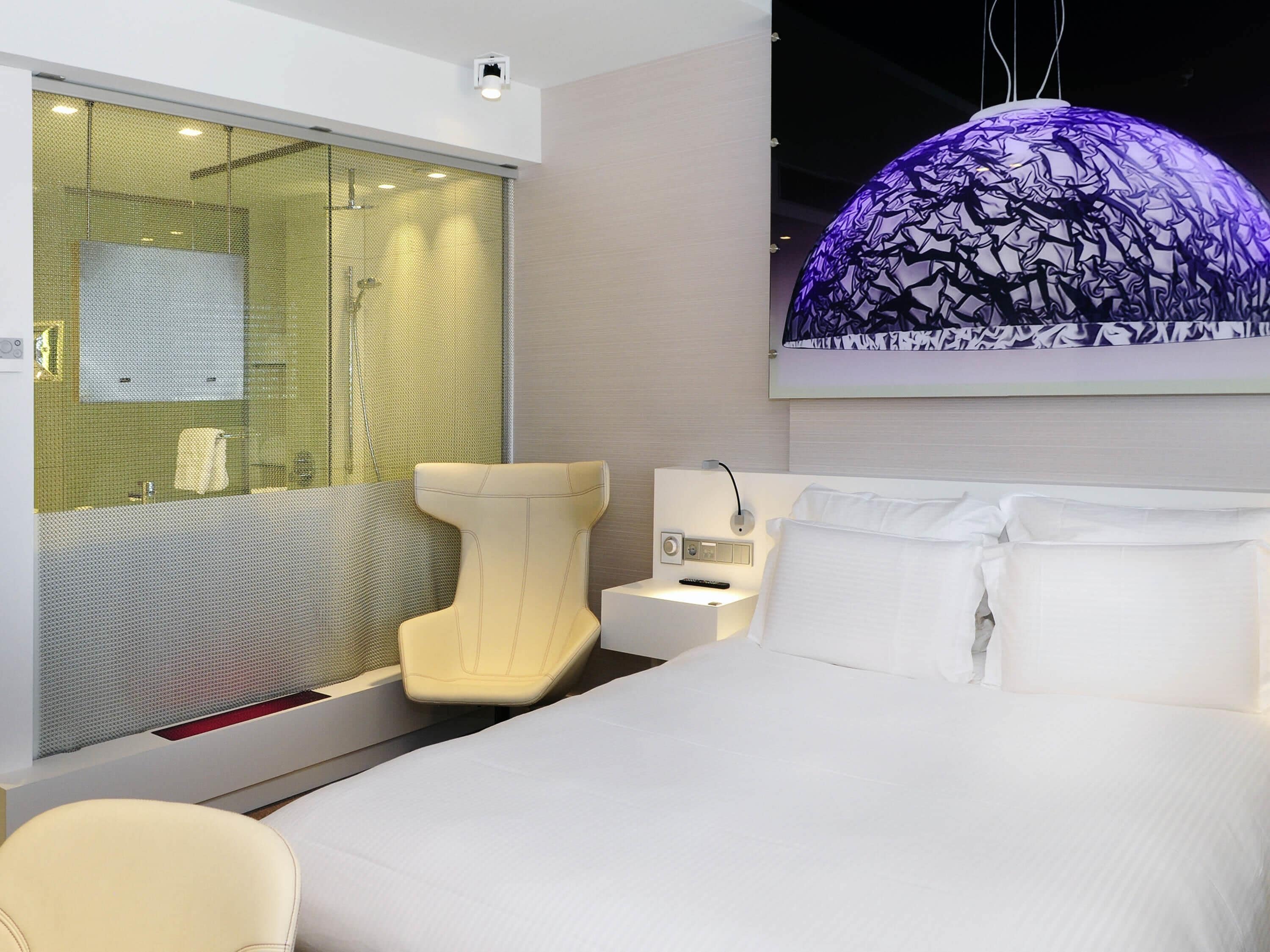 A modern hotel room with a bed, a chair, a desk, a bathroom with a shower, and a large window with a view.