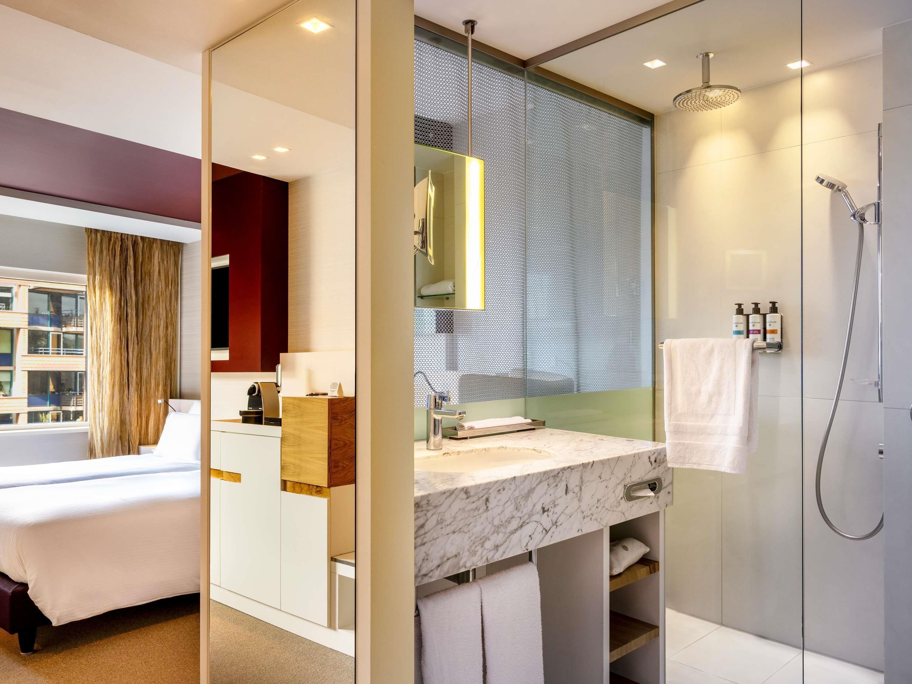 Una habitación de hotel moderna con una cama, un baño con ducha, un lavabo de mármol y un ventanal grande.